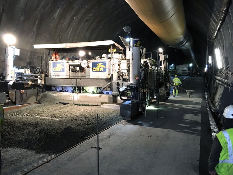 Photo: Tunnel construction site at which a slip-form paver is distributing concrete on the ground.