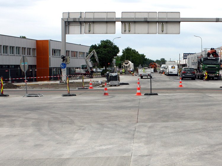 Foto: Frisch betonierte Fläche einer Kreuzung; im Hintergrund Baufahrzeuge, Warnkegel und Gebäude eines Gewerbegebiets