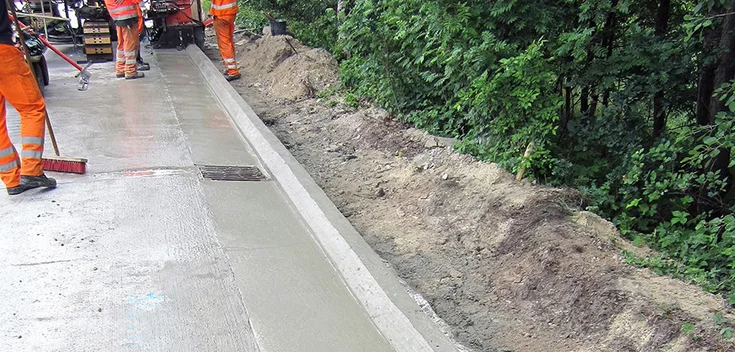 Foto: Frisch betonierte Straßenkante mit feucht glänzender Oberfläche; im Hintergrund Arbeiter und Baugeräte