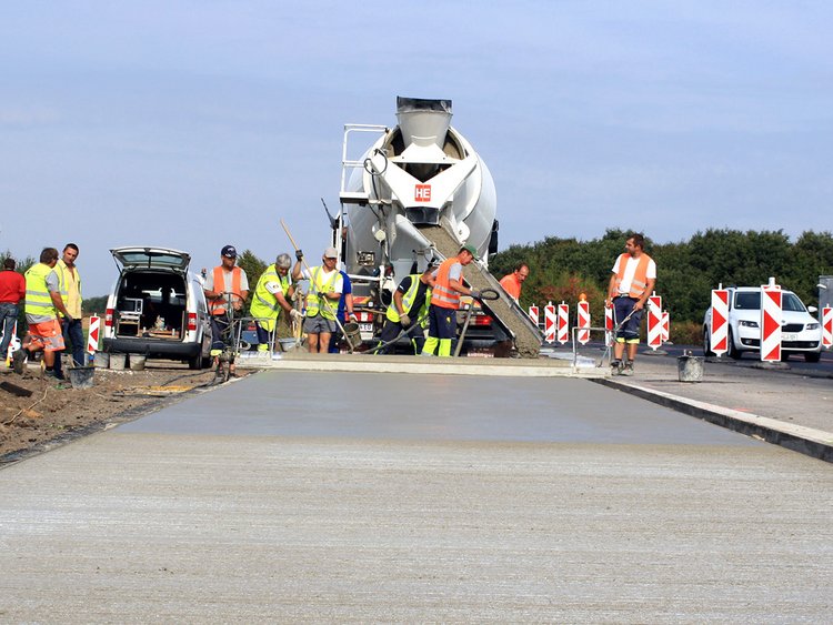 Foto: Frisch betonierte Fahrbahn; im Hintergrund zahlreiche geschäftige Arbeiter hinter einem Fahrmischer, aus dem flüssiger Beton auf den Boden geleitet wird