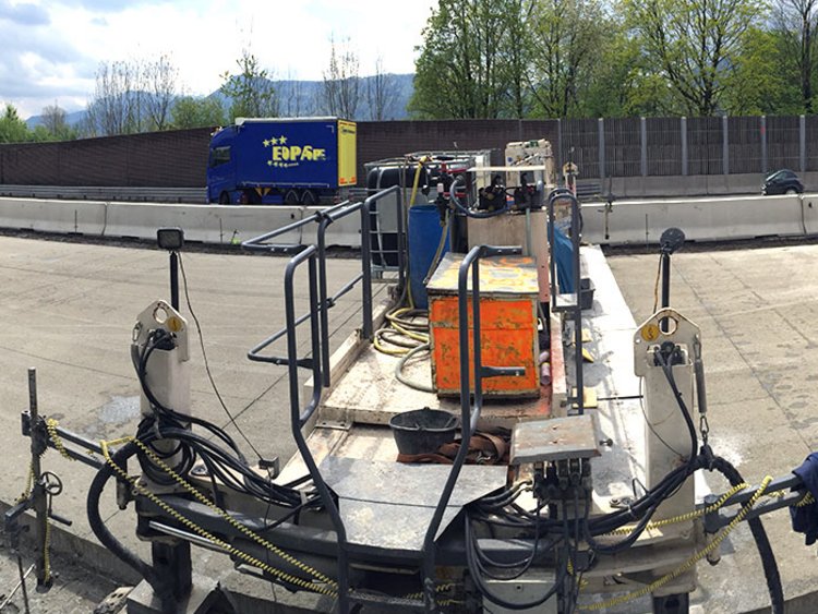 Foto: Panoramaaufnahme einer Straßenbaustelle; Betonfertiger und Arbeiter im Einsatz; im Hintergrund Bäume vor Berglandschaft