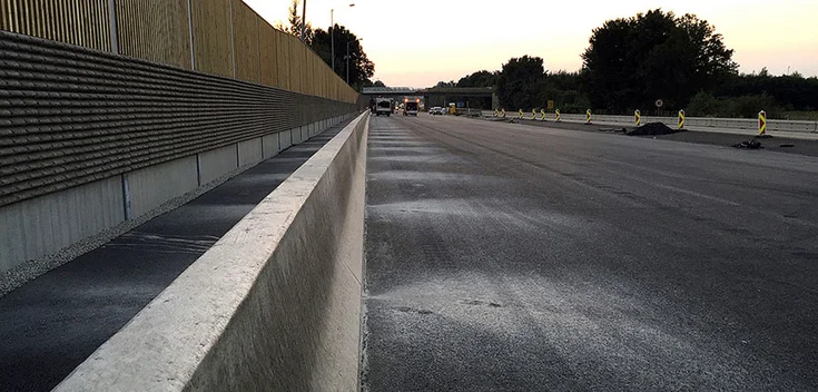 Foto: Abendaufnahme einer spiegelglatten Betonwand und Baustelleneinrichtung; im Hintergrund eine Brücke vor untergehender Sonne