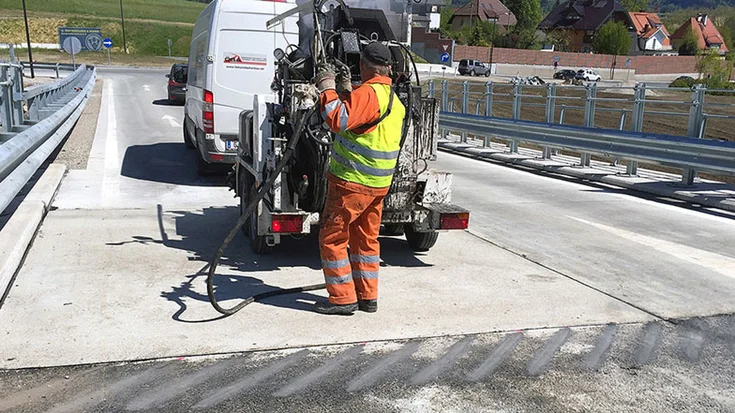 Foto: Baufahrzeug und ein Arbeiter, der am deutlich sichtbaren Übergang zwischen einer Beton- und einer Asphaltdecke Einstellungen an einer Baumaschine vornimmt