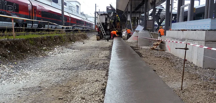 Foto: Gegossener, gegenüber dem mit Schotter bedeckten Untergrund erhöhter Betonstreifen; im Hintergrund Baugeräte und Arbeiter; links eine Personenzuggarnitur, rechts ein überdachter Bahnsteig