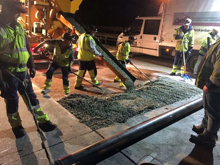 Photo: Night shot and rear view of a concrete mixer from which liquid concrete is flowing from a pipe; two workers distribute it evenly on the ground; other workers observe the scene.