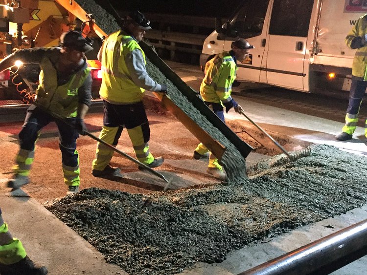 Foto: Nachtaufnahme und Rückansicht eines Betonmischers, aus dem über eine Rohleitung flüssiger Beton fließt; zwei Arbeiter verteilen ihn gleichmäßig am Boden; weitere Arbeiter beobachten die Szene.