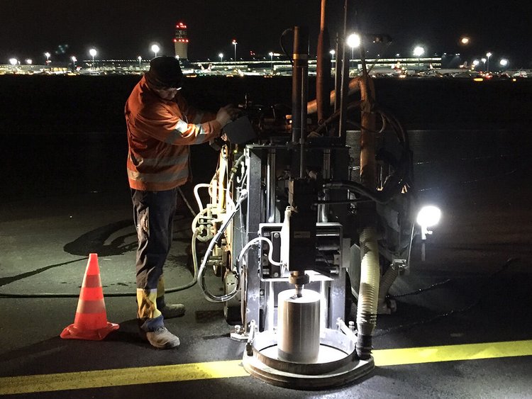 Foto: Asphaltfläche mit gelber Markierung bei Nacht; ein Arbeiter mit Bohrgerät; im Hintergrund hell erleuchteter Flughafen
