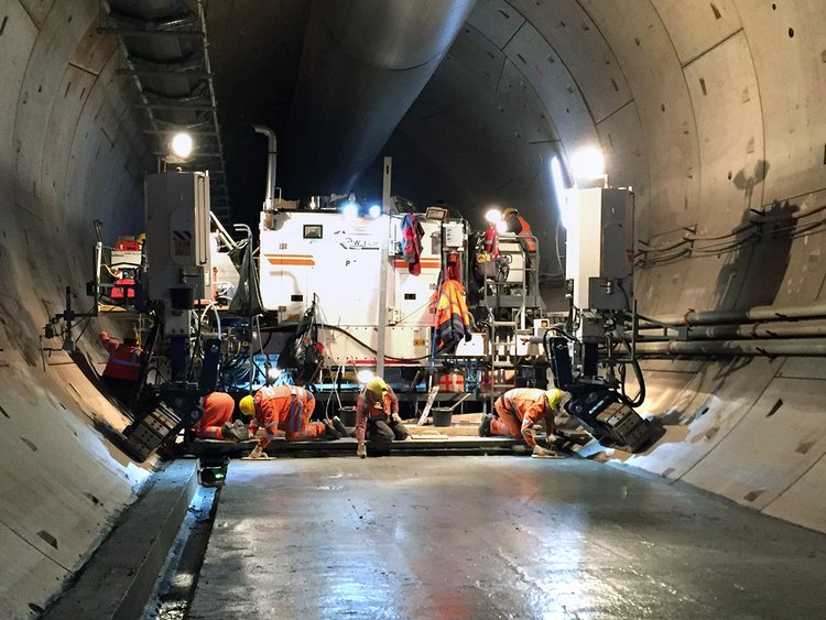 Foto: Glatte, glänzende Betondecke in einer Tunnelröhre; im Hintergrund der Gleitschalungsfertiger, auf dessen hinterer Plattform vier Arbeiter an der Betonoberfläche arbeiten