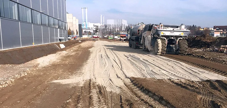 Foto: Aufgewühlte Erdfläche mit zahlreichen Reifenspuren; links eine Industriehalle mit silbergrauer Verkleidung; rechts Baufahrzeuge mit überdimensioniert wirkenden Reifen