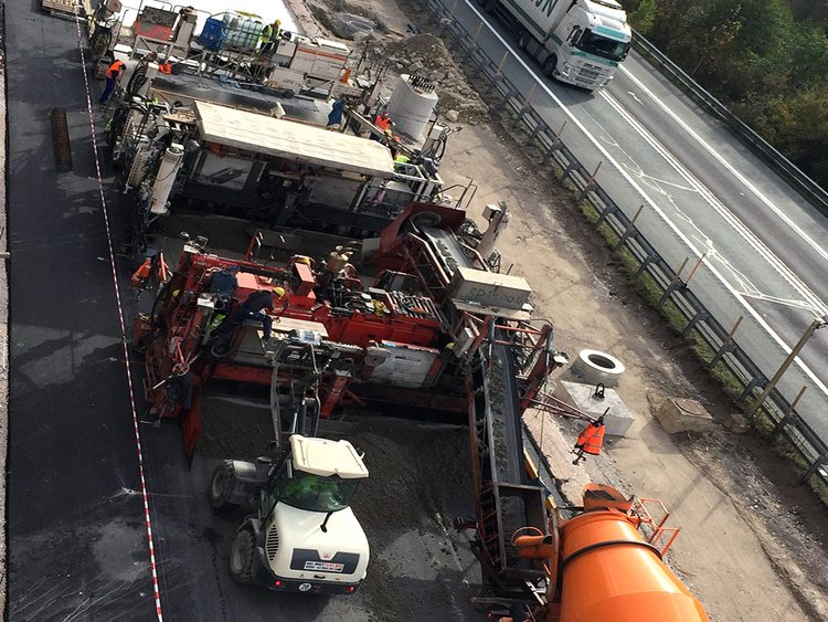 Foto: Luftaufnahme einer Straßenbaustelle; Einbauzug beim Einbau einer frisch betonierten Fahrbahn; Lkw-Verkehr auf Gegenfahrbahn