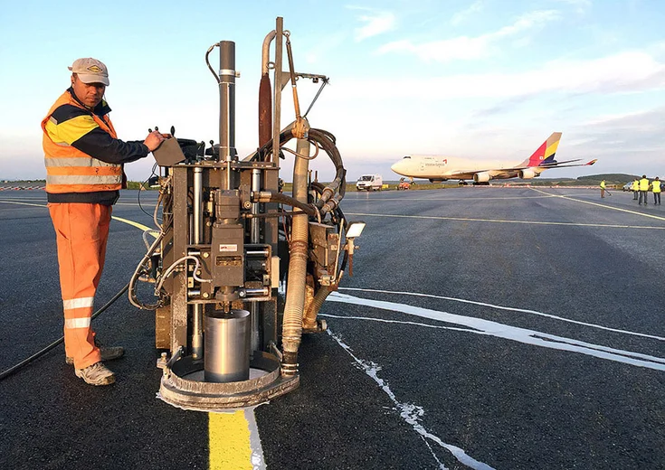 Foto: Arbeiter am Bedienpult einer Maschine auf einem Flugfeld; im Hintergrund Flughafenpersonal und eine Boeing 747