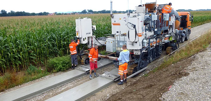 Foto: Zwei frisch gegossene, gegenüber dem erdigen Untergrund erhöhte Betonstreifen; ein komplexes Baufahrzeug und konzentriert tätige Arbeiter