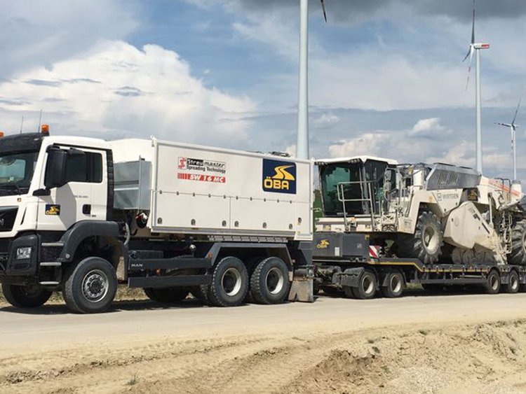 Foto: Lkw-Zug mit großer Baumaschine auf einem Tieflader auf einer fest gewalzten Sandstraße; im Hintergrund Grünland und Windräder 