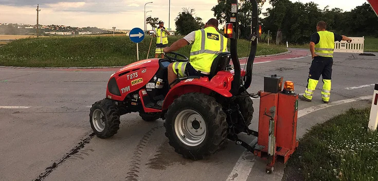 Foto: Drei Arbeiter mit einem Vermessungsgerät und einer Baumaschine am Rand eines Kreisverkehrs
