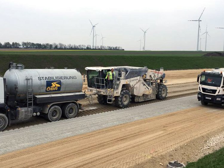 Foto: Ebenmäßig gestrichener, heller Sand auf einem Straßenverlauf; darauf Transport- und Spezialbaufahrzeuge; Im Hintergrund grünes Flachland und Windräder