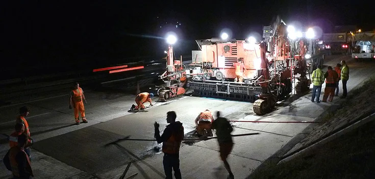 Foto: Nachtaufnahme einer Baustelle mit einer teils bereits aufgetragenen Betonschicht, zahlreichen Arbeitern und einer großen Baumaschine mit Scheinwerfern, die die Baustelle in helles Licht hüllen